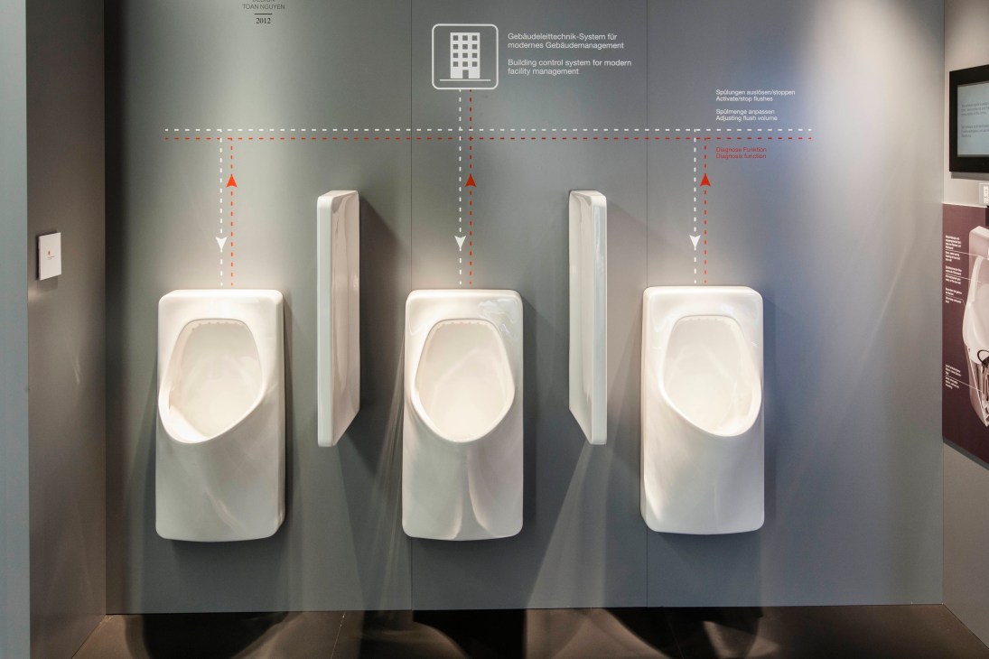 Three white wall-mounted urinals displayed at a trade show with schematic diagrams above.