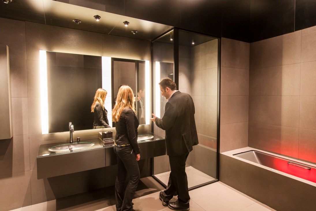 Two professionals examining a modern bathroom display with illuminated mirrors and sleek fixtures at a trade fair.