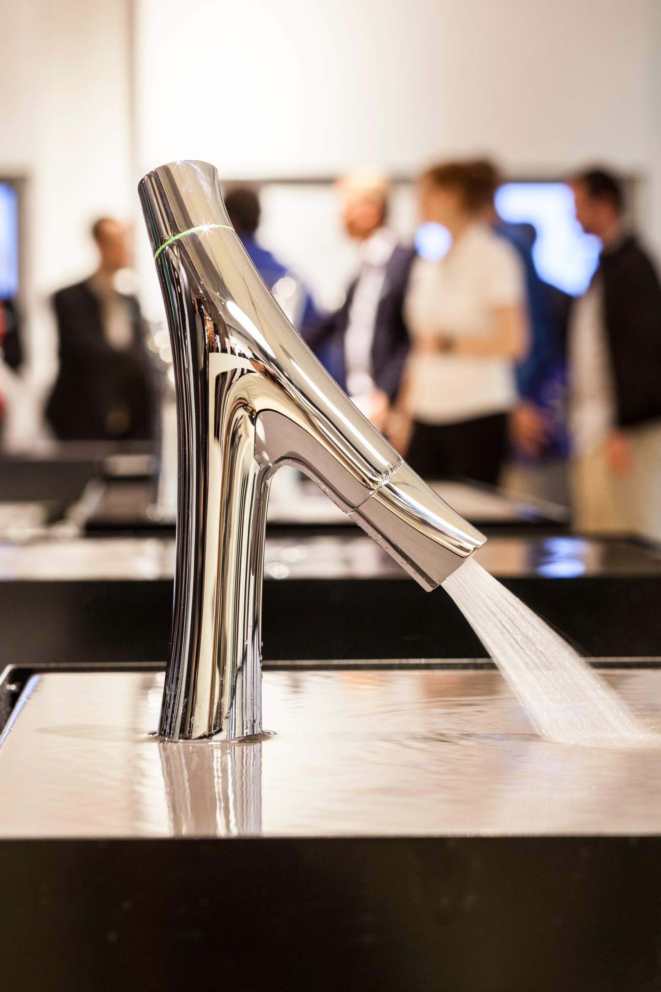 Chrome tap from the Axor Starck line with water flowing, displayed at a trade fair with visitors in background.