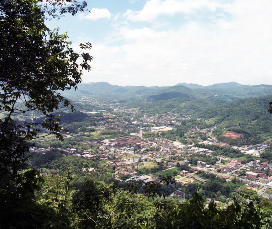Aerial view of Pomerode valley with forested mountains, organized streets, and buildings nestled in tropical landscape.