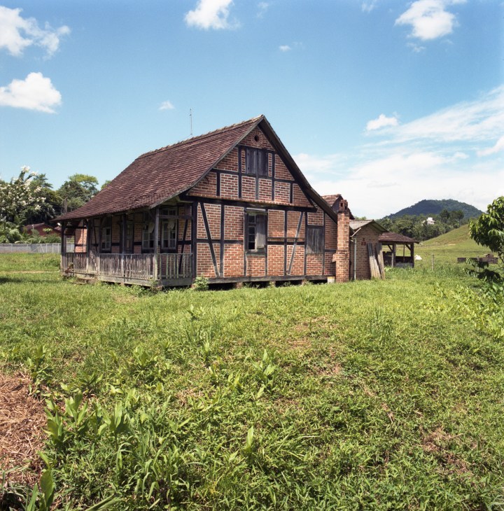Traditional half-timbered brick house with pitched roof in Pomerode, Brazil, surrounded by cultivated fields and mountains.