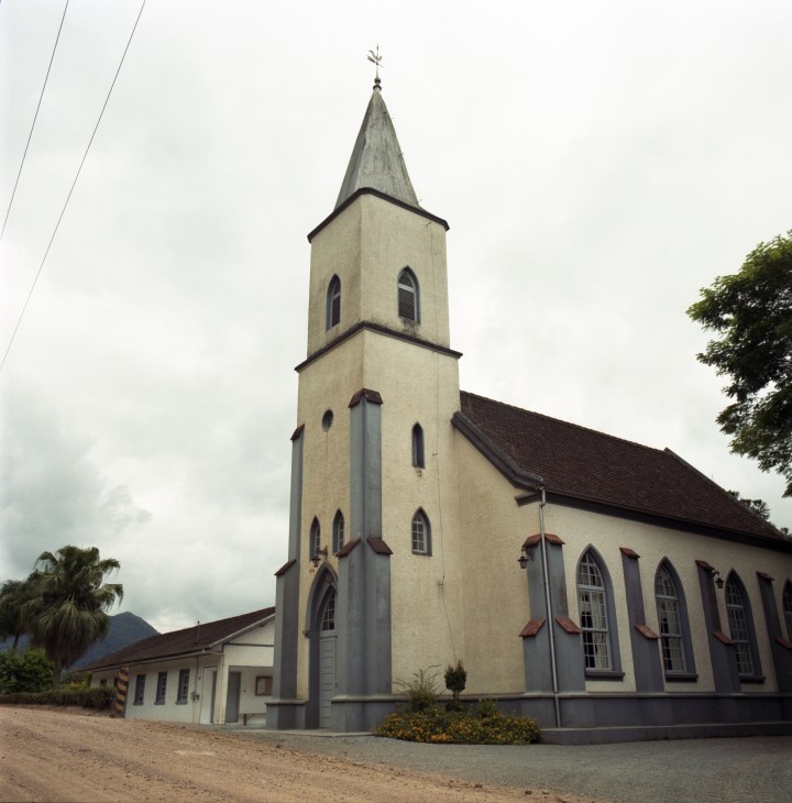 A cream-colored Lutheran church with a tall pointed steeple and cross, featuring arched windows and dark roof trim, with palm trees visible in the background.