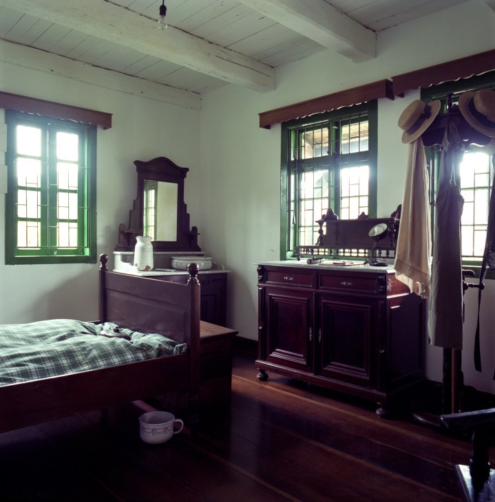 A traditional bedroom with dark wooden furniture, white walls, green-framed windows and period furnishings at Casa do Imigrante in Pomerode.