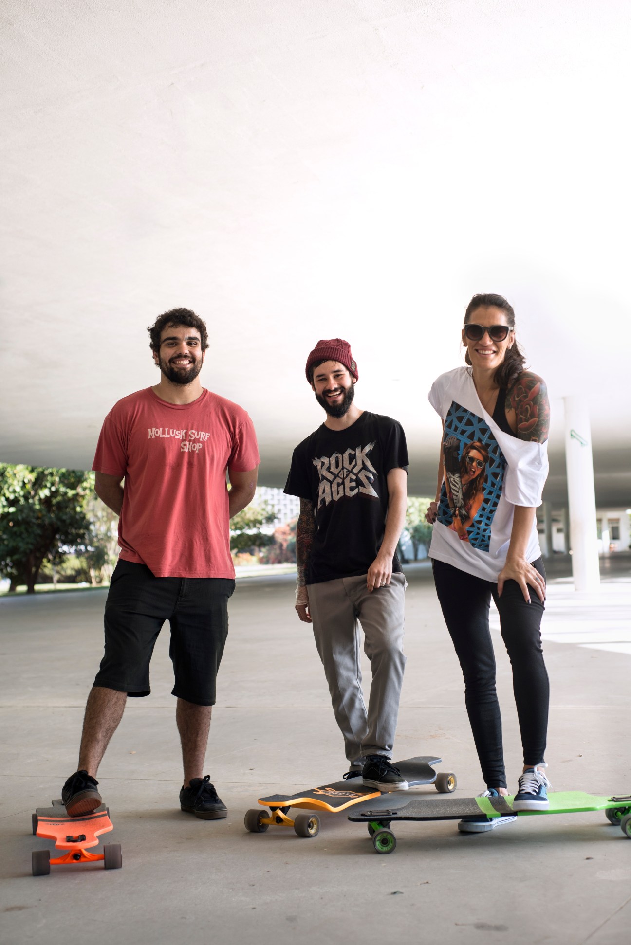 Three people with skateboards standing on a concrete plaza with green landscape and buildings in background.