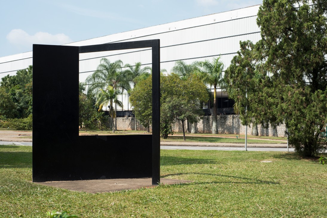 Black minimalist frame sculpture in grassy area of Ibirapuera Park with palm trees and institutional buildings visible in background.