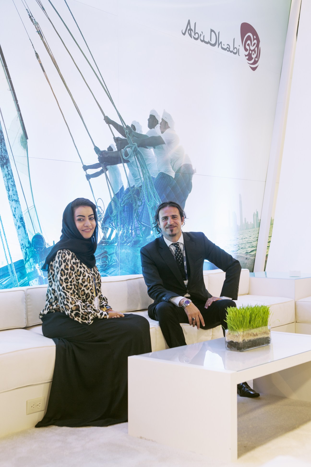 Two people seated at an Abu Dhabi cruise terminal exhibition booth with sailing imagery backdrop.