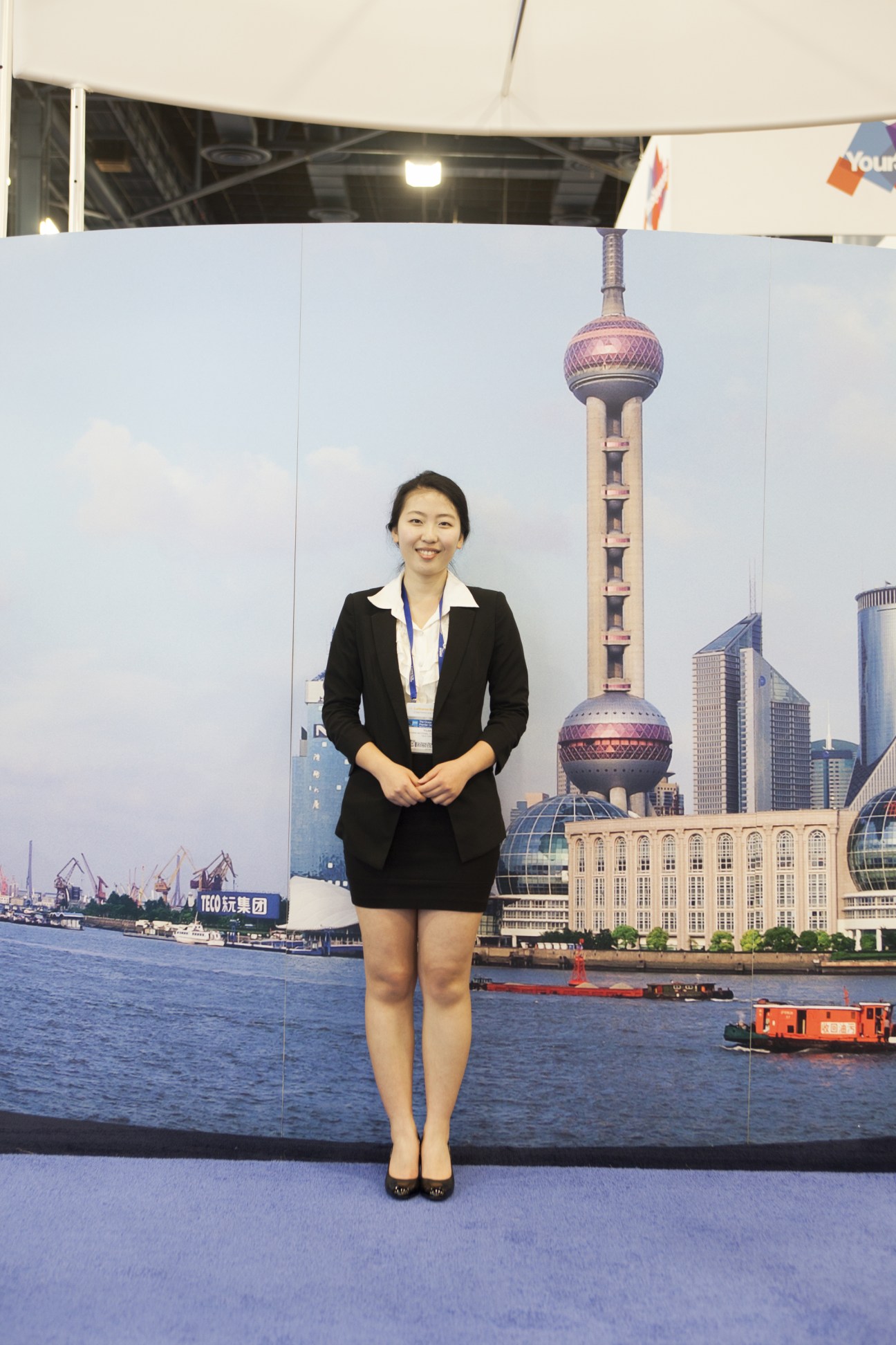 Woman in black business suit standing in front of Shanghai skyline backdrop at trade show.