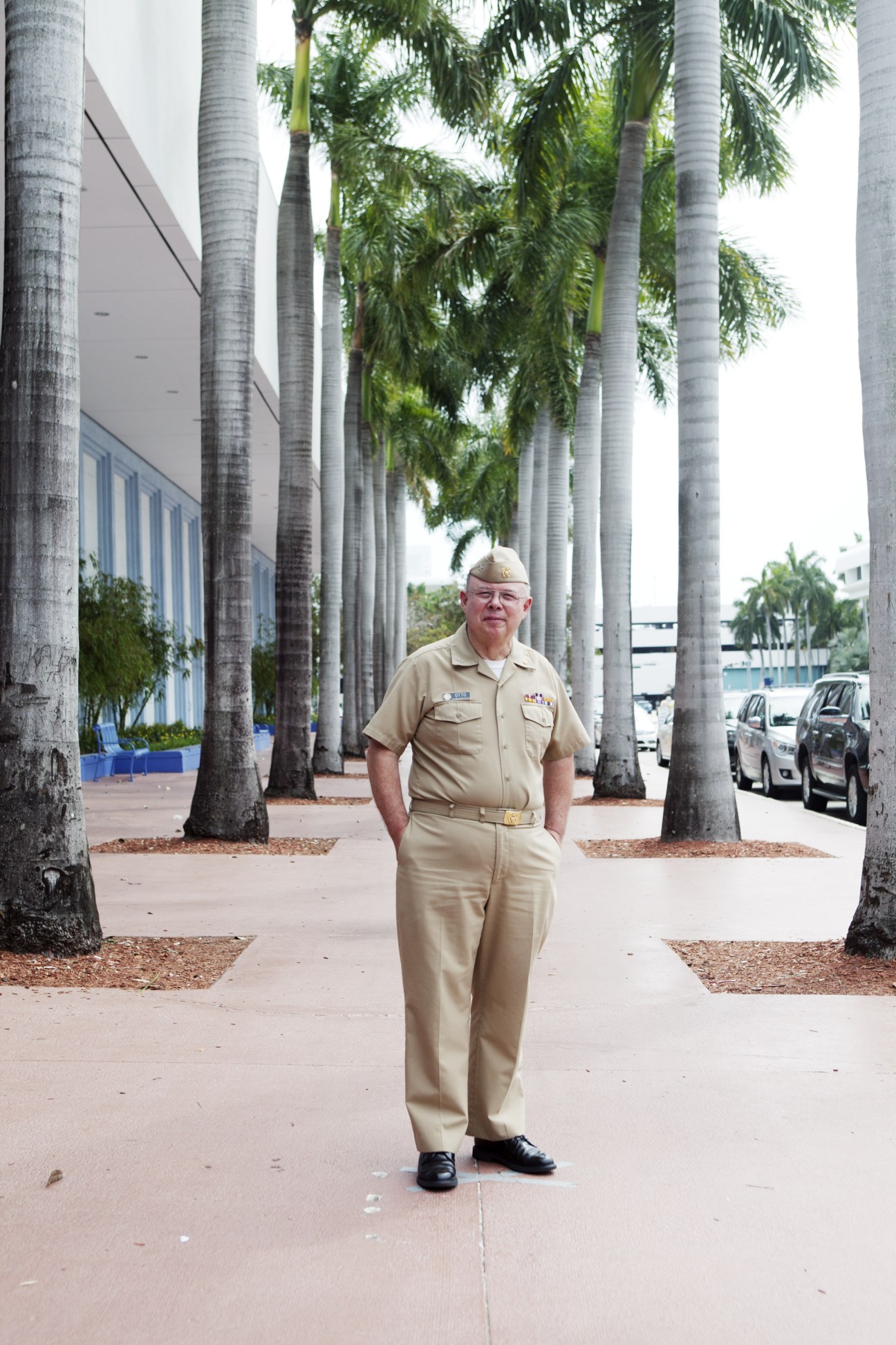 Military officer in tan uniform standing under palm trees at a cruise terminal.