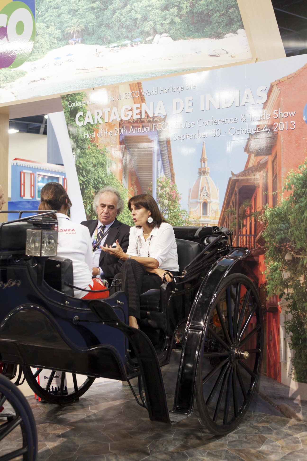 Two people sit in a traditional black horse-drawn carriage at a Cartagena cruise conference exhibition booth decorated with colonial architecture imagery.