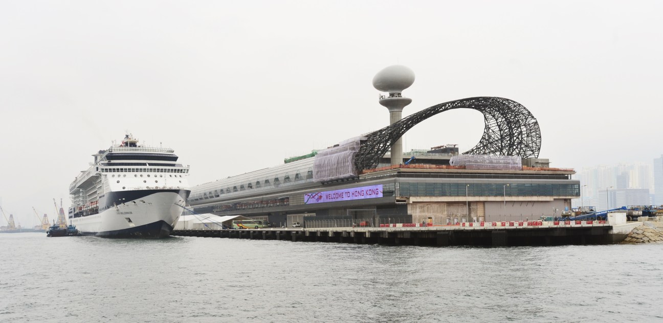 Large cruise ship docked at Hong Kong's new cruise terminal with distinctive curved architectural structure.