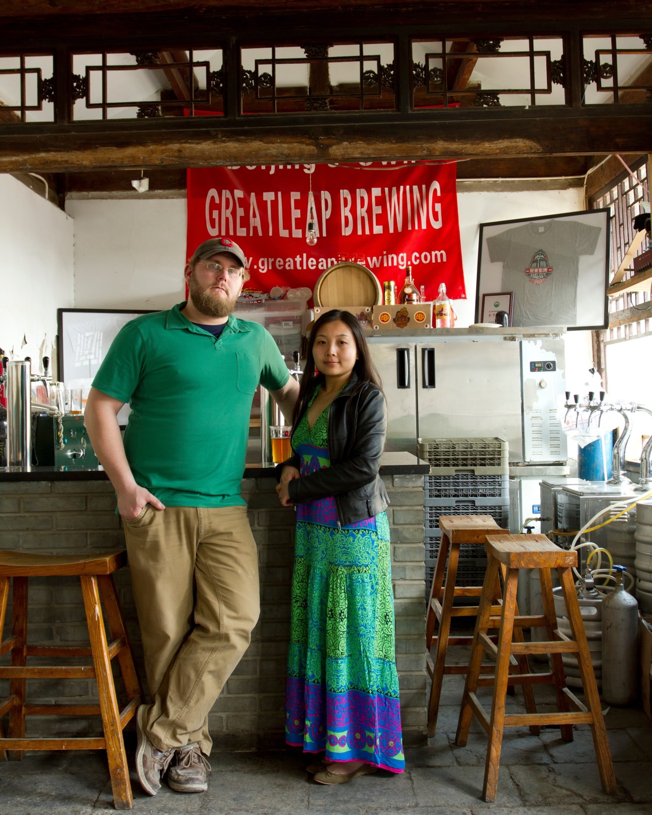 Carl Setzer and Liu Fang stand in Great Leap Brewery's interior with red signage and brewing equipment visible behind them.