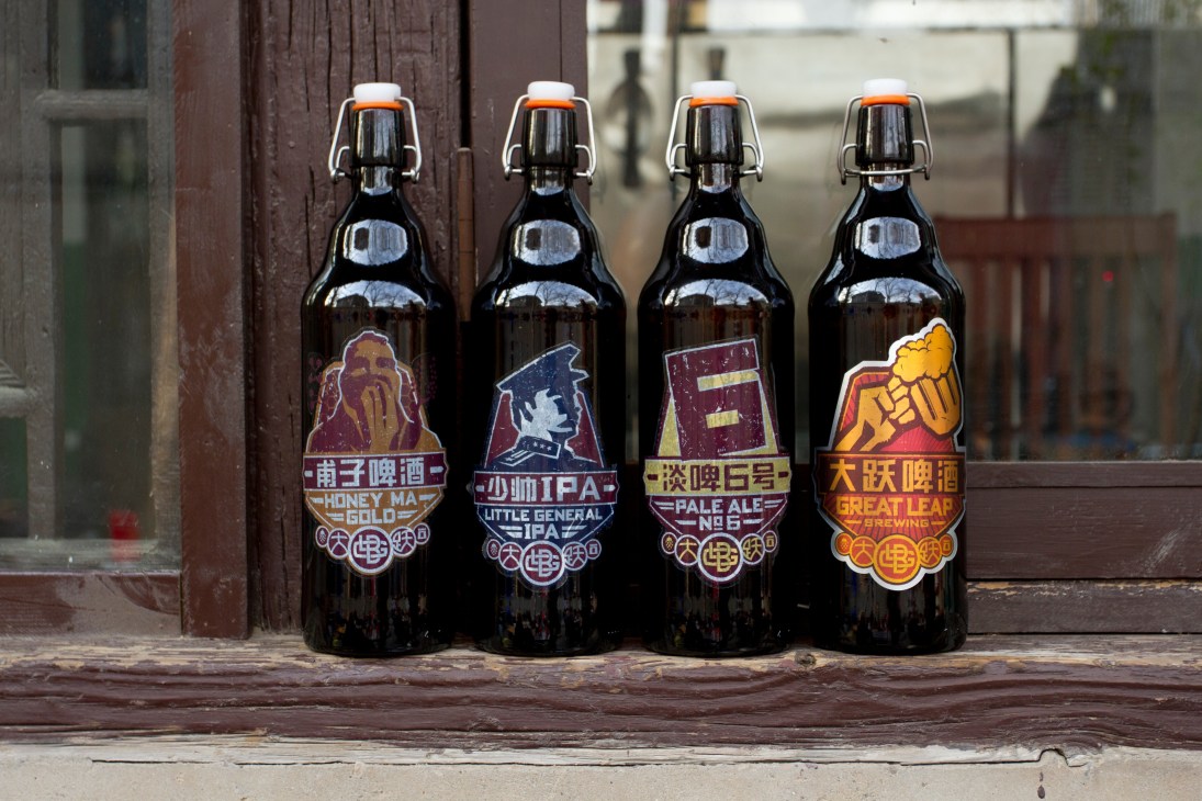 Four Great Leap Brewery beer bottles with colorful labels featuring Chinese characters and English text displayed against a brick wall.