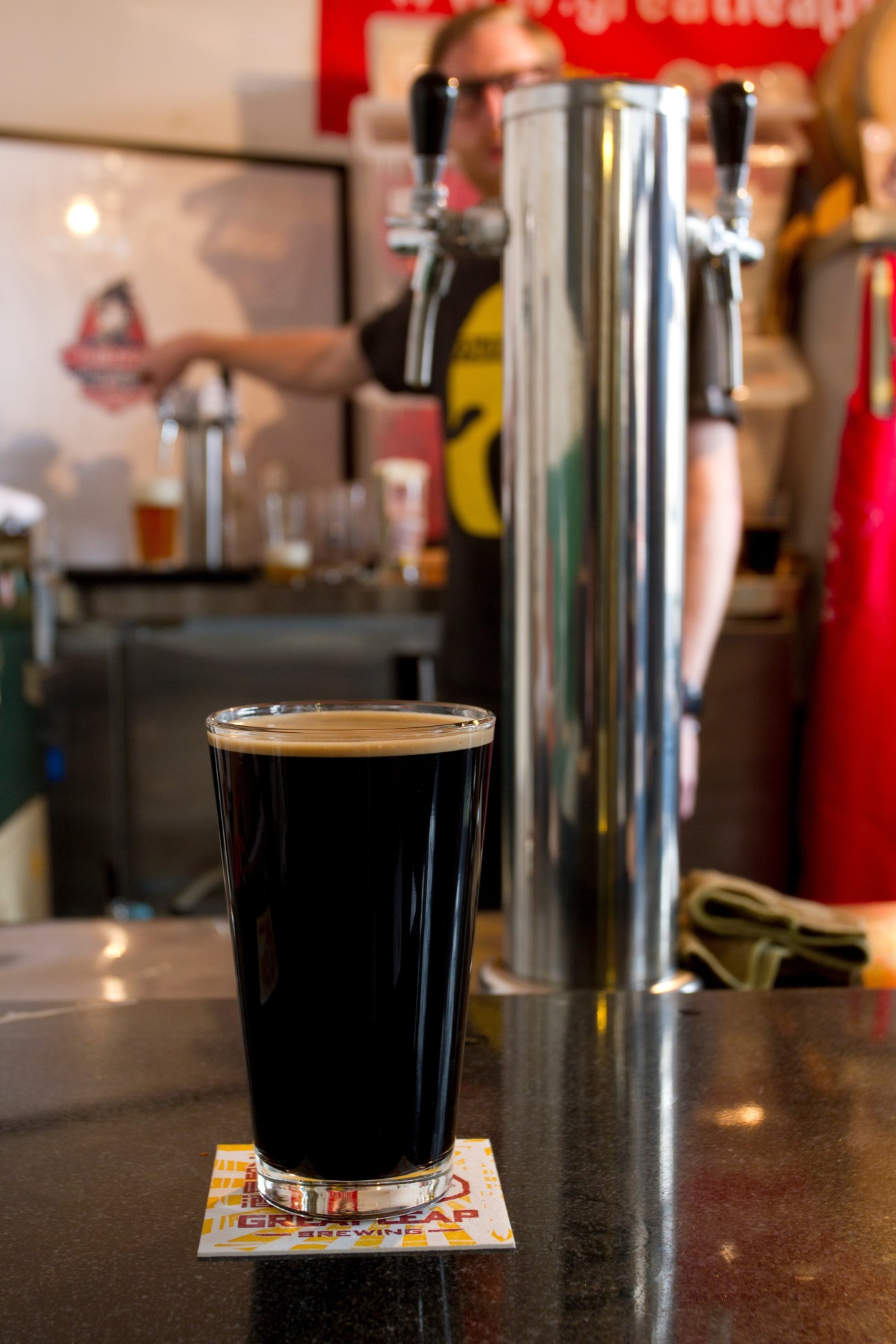 A glass of dark beer sits on a bar counter with beer taps and brewery staff blurred in the background.