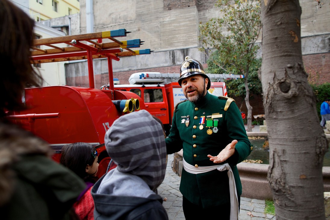 A Chilean firefighter in green uniform with medals explains fire safety to children next to a red fire truck.