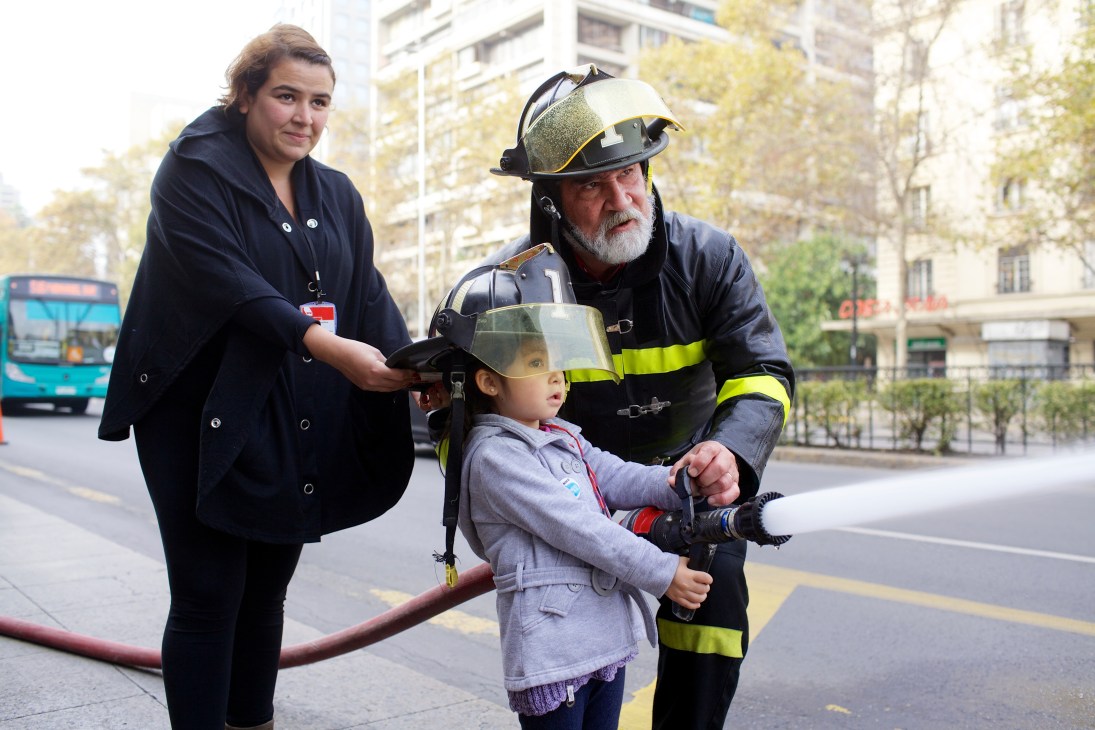 Firefighter and woman instructing a child on using firefighting equipment during a public demonstration in Santiago.