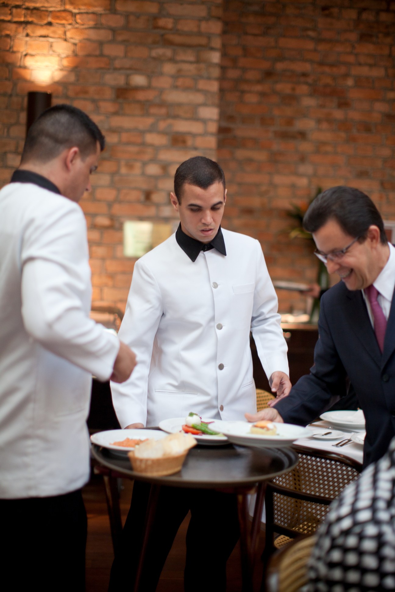 Service staff in white and formal attire attending to diners at a restaurant table.
