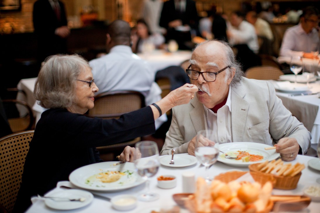 An elderly man and woman dining together at a restaurant table, sharing food from each other's plates.
