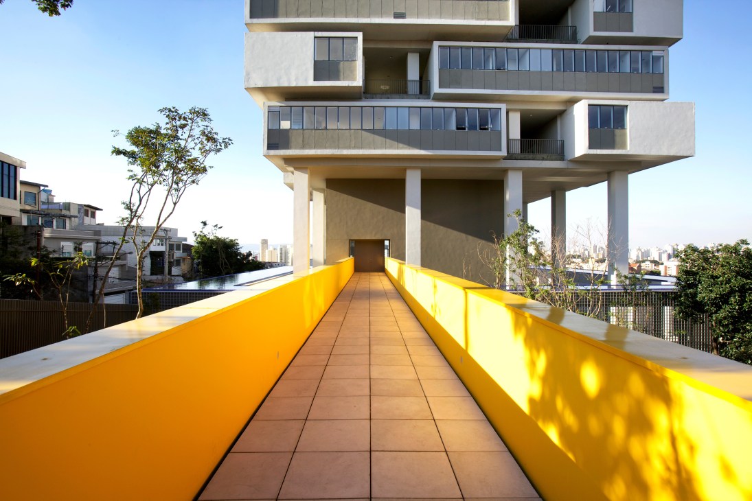 Yellow suspended walkway leading to the 360º Building entrance with stacked cuboid apartments above and São Paulo skyline beyond.