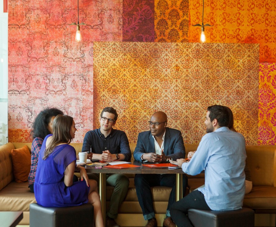 Five people in business casual attire sit around a table having a discussion in a modern office space with colorful patterned wallpaper.
