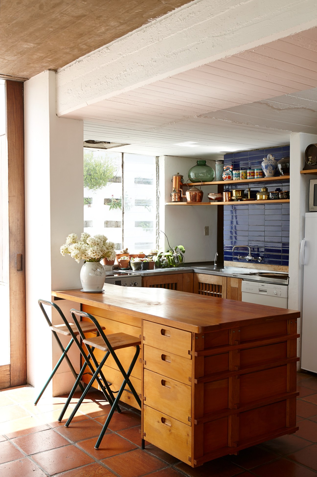 Die Es kitchen with wooden island, blue tiled backsplash, open shelving and natural light from large windows.