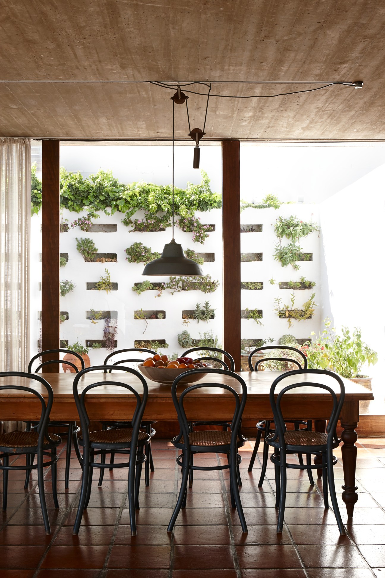 Die Es dining area with wooden table, black chairs, concrete ceiling with track lighting, and living wall of plants beyond glass doors.