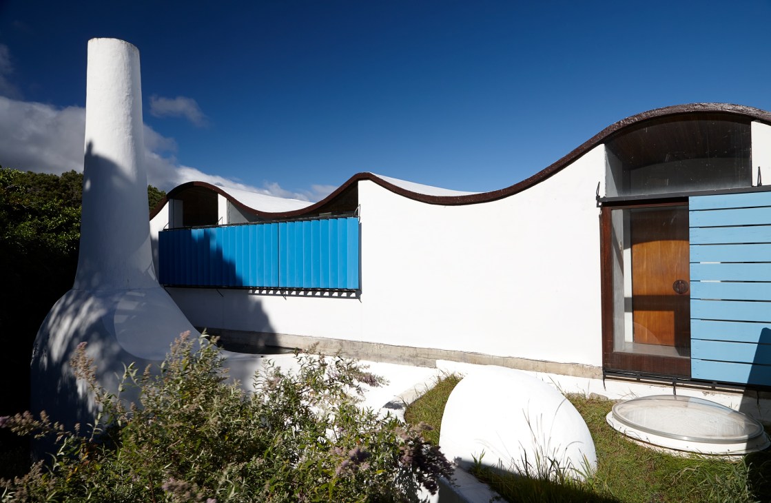 White modernist architecture with wavy roof line, cylindrical chimney, blue railing and wooden door in landscape setting.