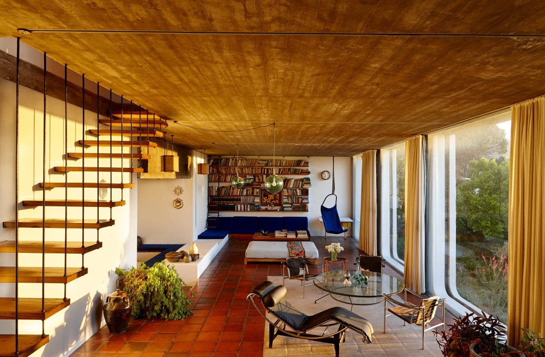 Living room with concrete ceiling, wooden staircase, bookshelves, and large windows overlooking landscape.