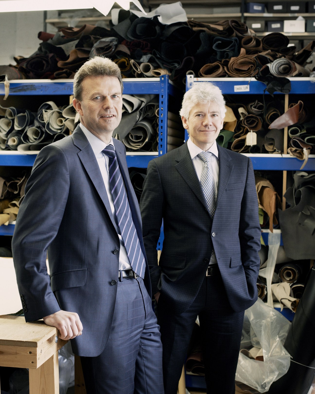 Two men in business suits stand in front of shelves of rolled leather at Joseph Cheaney & Sons shoemaking factory.