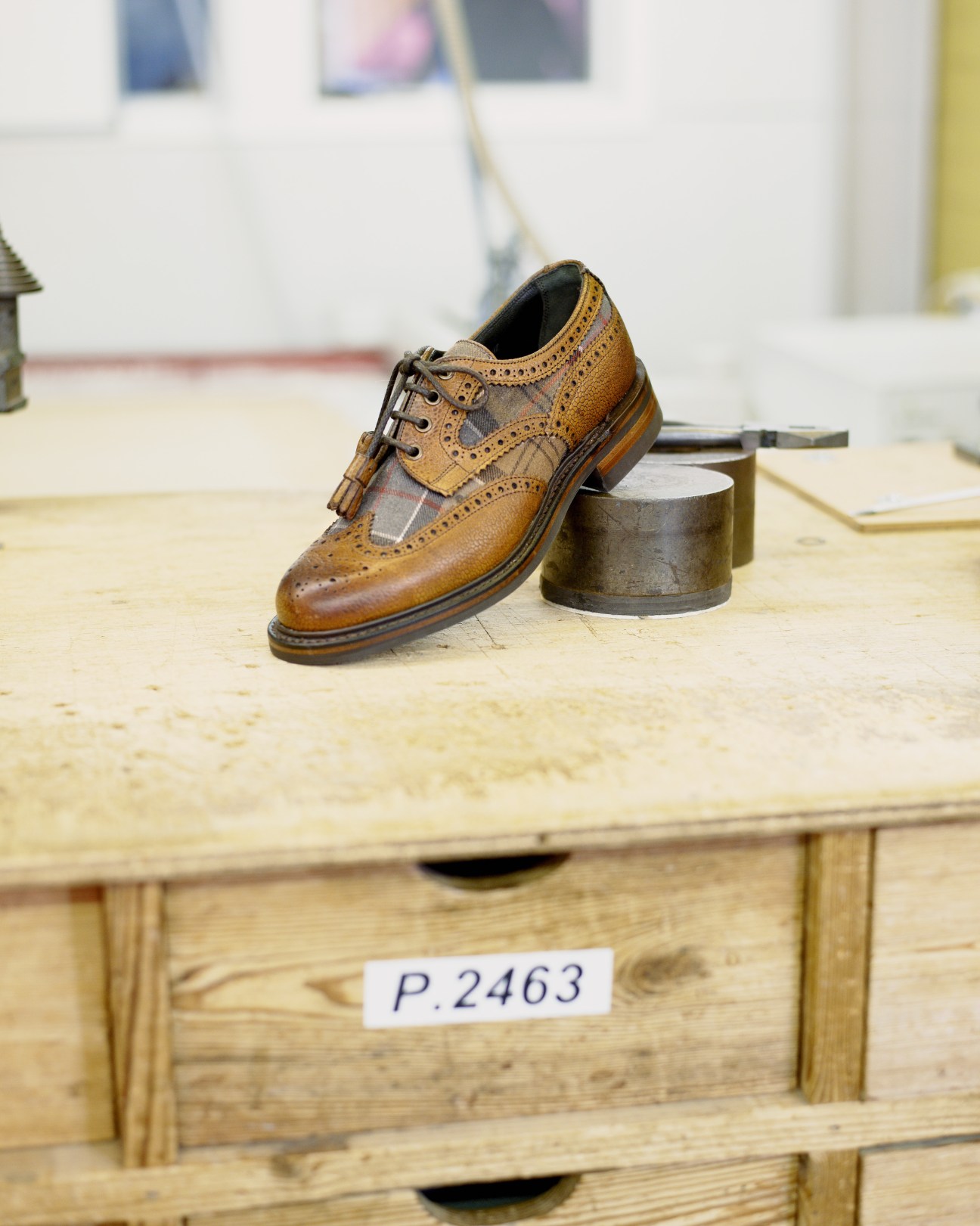 A tan and brown leather brogue shoe with detailed stitching displayed on a wooden workbench in a shoemaking factory.