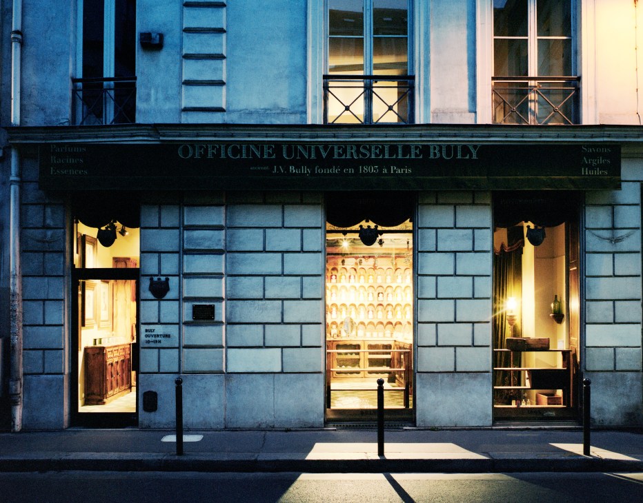 Storefront of Officine Universelle Buly pharmacy in Paris with illuminated windows displaying products on shelves.