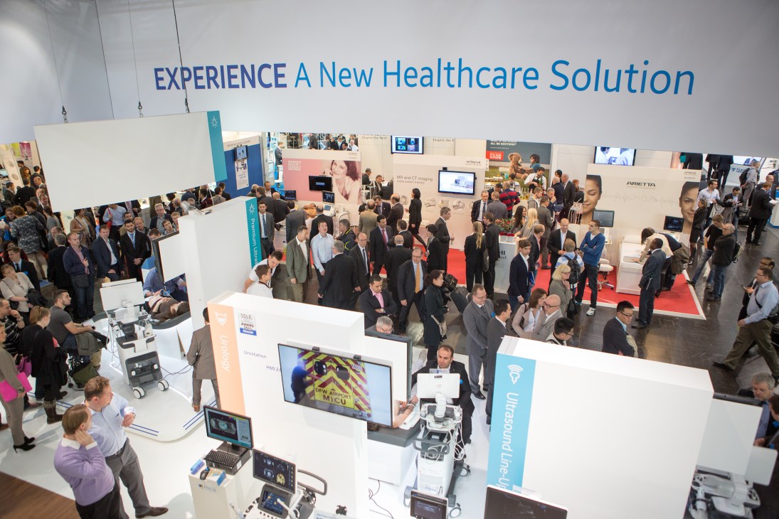 Overhead view of the Medica trade fair floor with numerous exhibitor booths, visitors, and signage reading 'EXPERIENCE A New Healthcare Solution'.