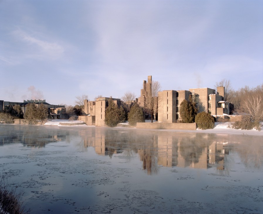 Champlain College at Trent University reflected in the frozen Otonabee River, with brutalist concrete structures and evergreen trees.