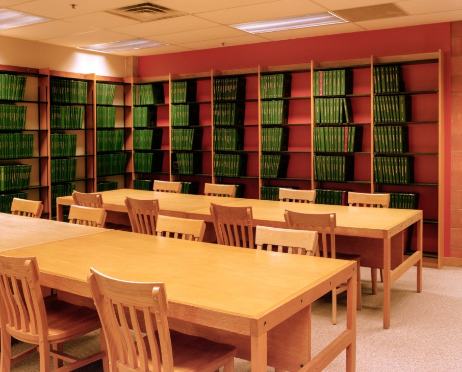 Library reading room with wooden tables, chairs, and red-painted shelving filled with green-bound books.