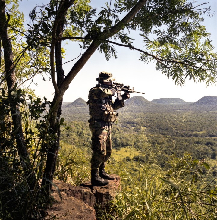 Armed Senad special forces member in camouflage standing on jungle terrain with rifle, surrounded by dense vegetation and mountains.