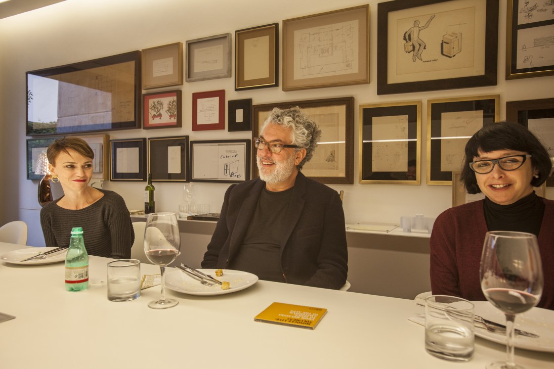 Marcio Kogan and Suzana Glogowski seated at a dining table in an office with framed architectural drawings on the wall behind them.
