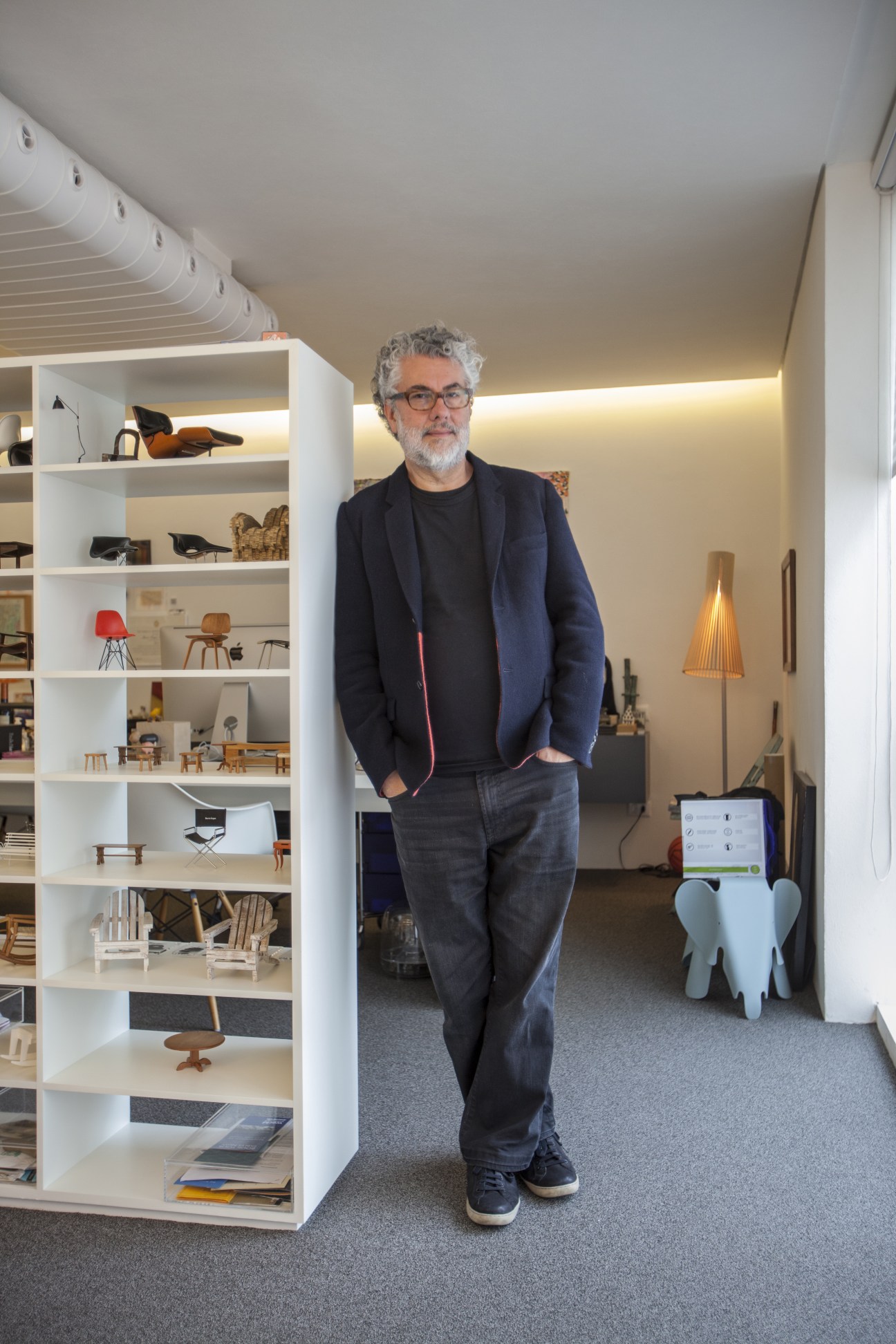 Marcio Kogan stands in his minimalist office featuring white shelving displaying design objects and furniture models.