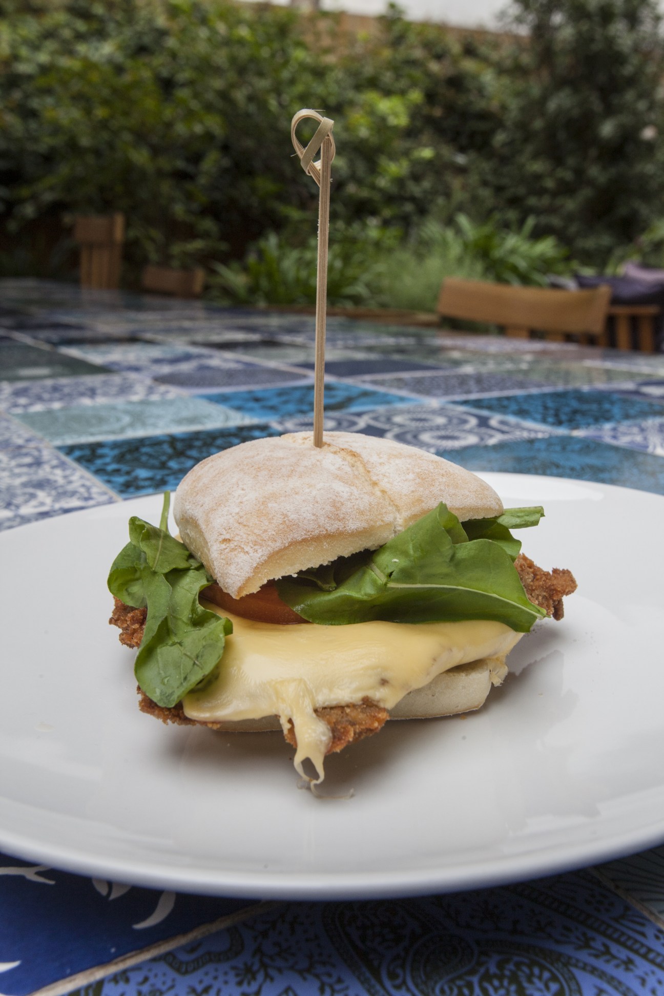 Milanesa sandwich with breaded beef, cheese, rocket and tomato on ciabatta bread with a pick, served on a white plate outdoors.
