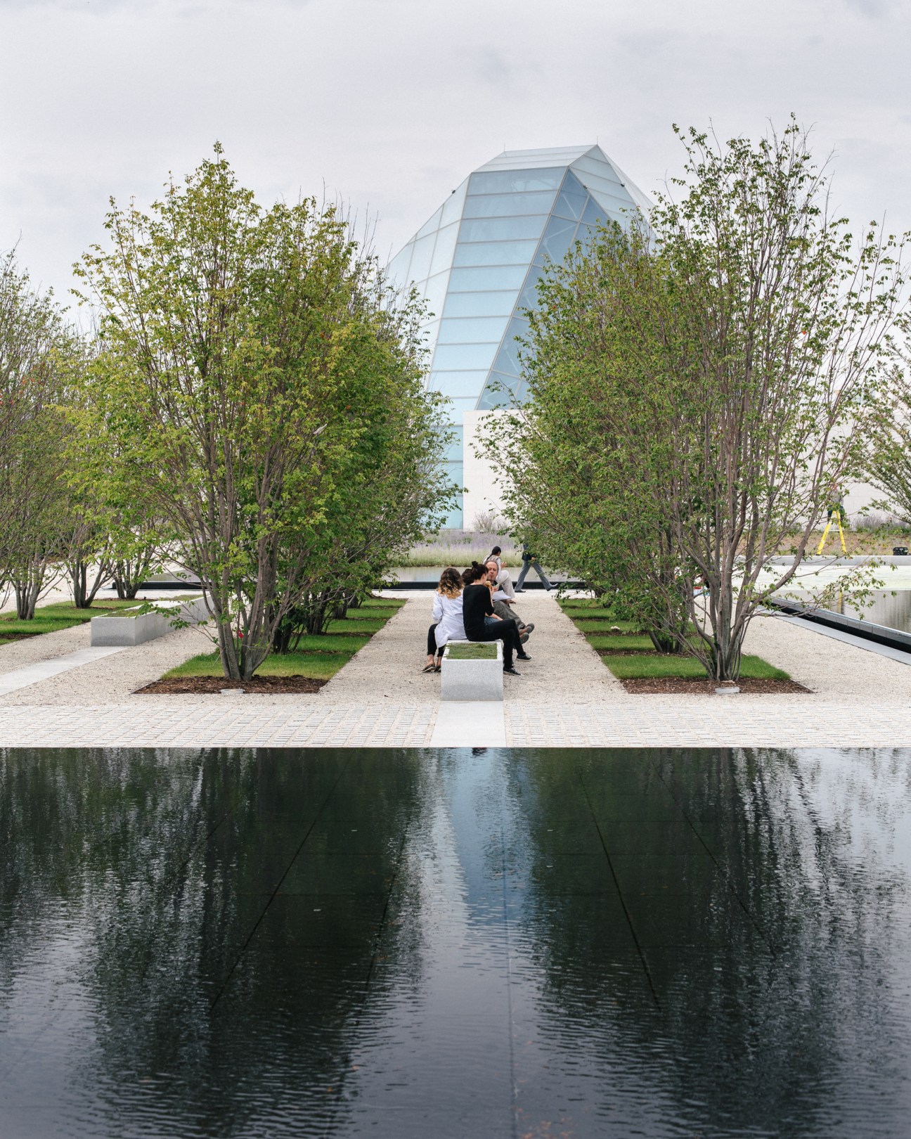 Visitors walk through an Islamic formal garden with rows of trees flanking a reflective pool, with the Ismaili Centre's glass pyramid visible in the background.