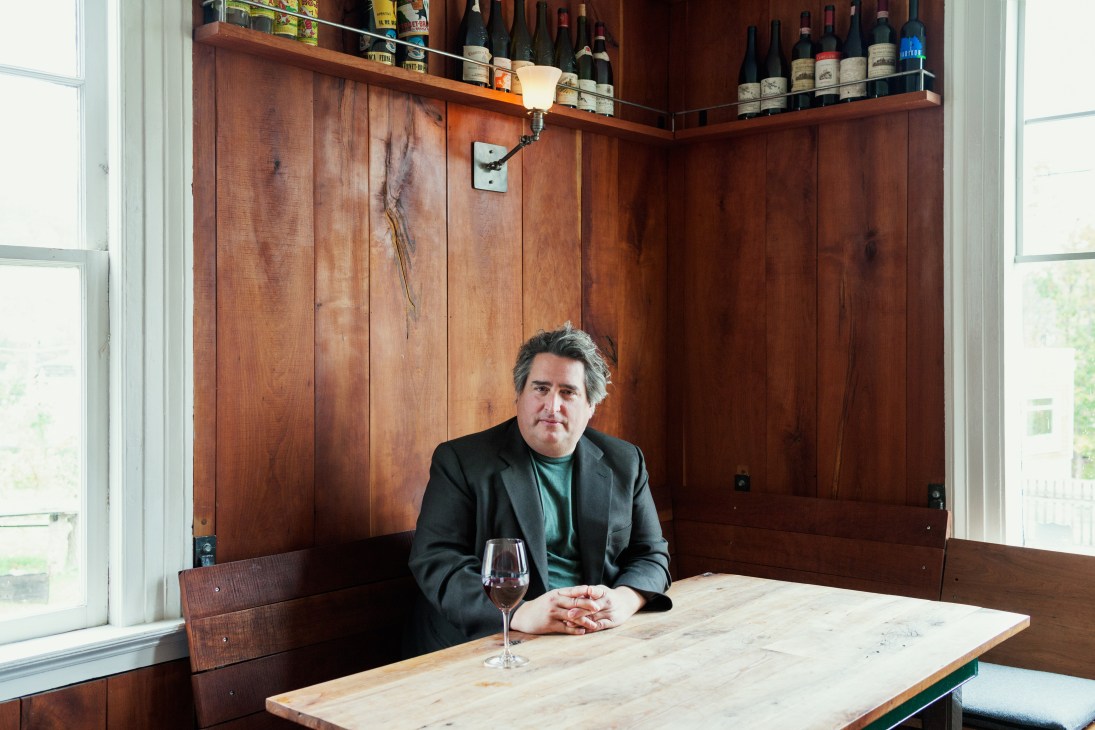 Photographer Gregory Crewdson seated at a wooden table with a wine glass, in front of wooden paneling and wine bottles.