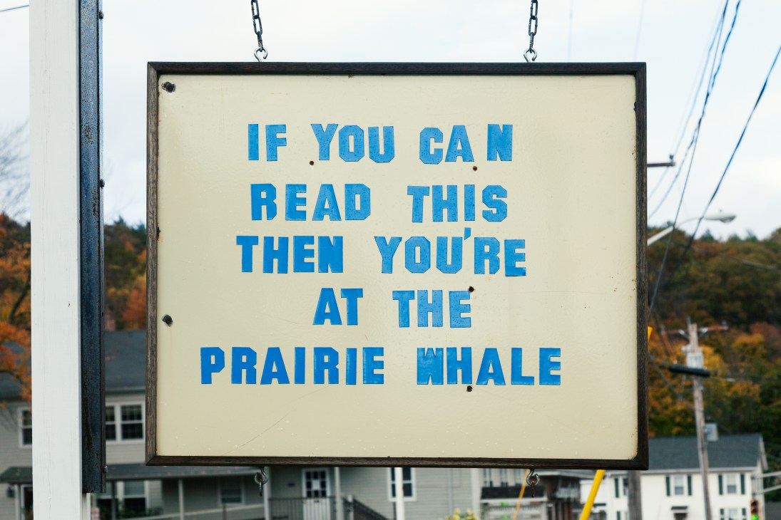 Weathered cream and blue sign reading 'If you can read this then you're at the Prairie Whale' hanging from chains with autumn foliage visible behind.