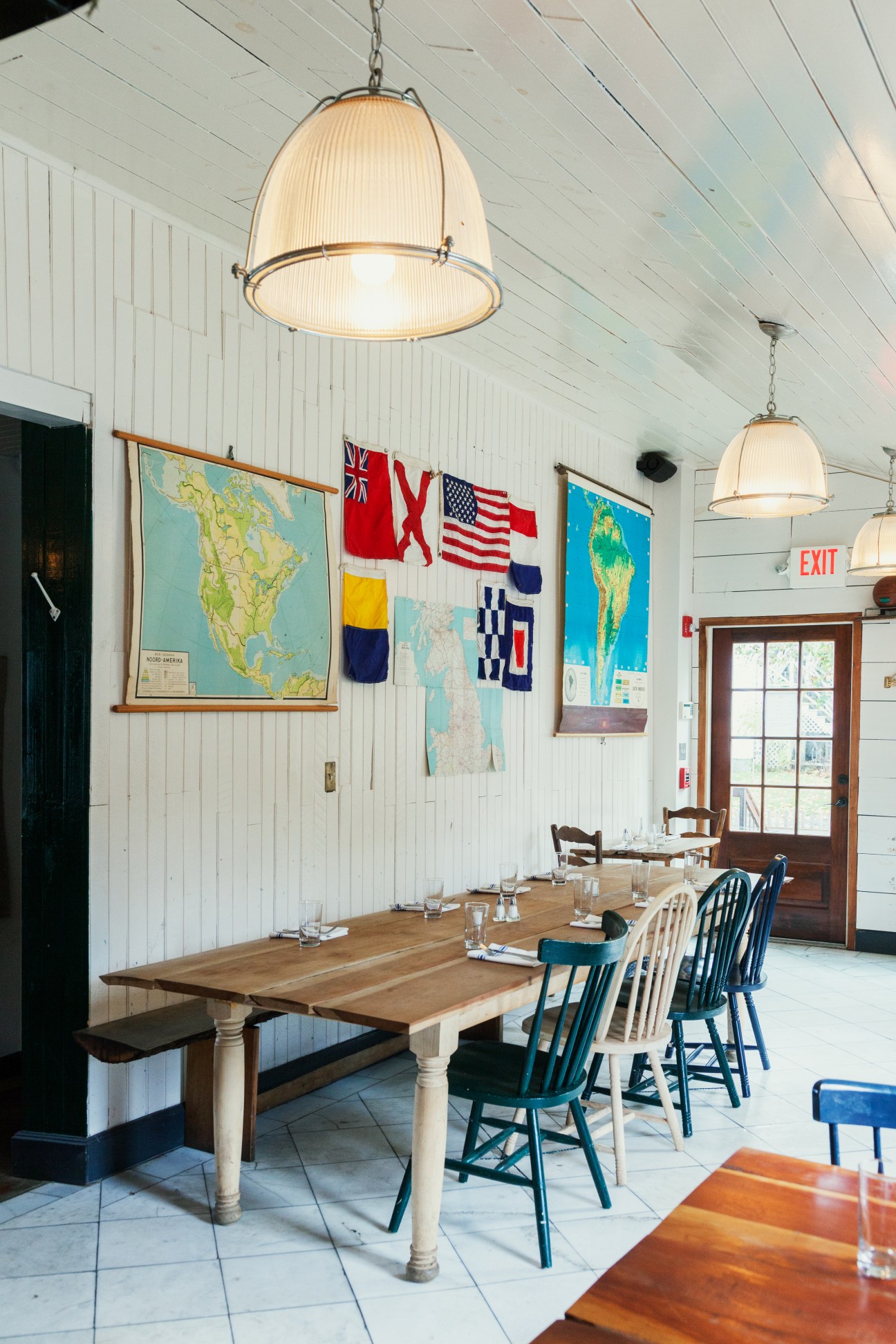 Prairie Whale restaurant's front dining area with long wooden table, Windsor chairs, and vintage maps on white shiplap walls.