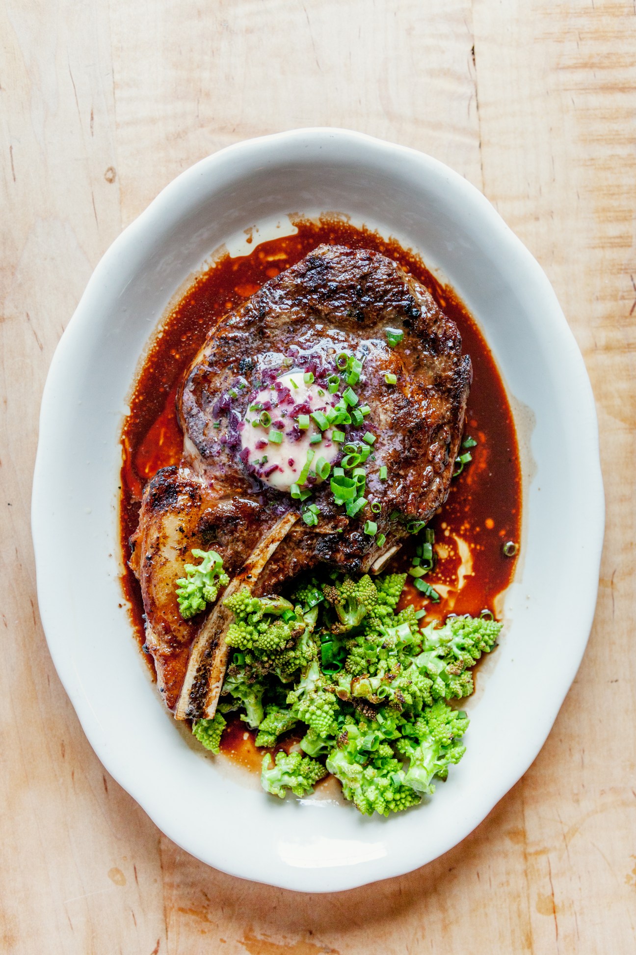 Rib-eye steak on the bone with romanesco cauliflower, garnished with scallions and sour cream on a white plate.
