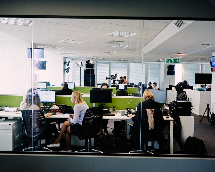 Open-plan office space with staff working at desks with green dividers and computer monitors, with filming equipment visible in the background.