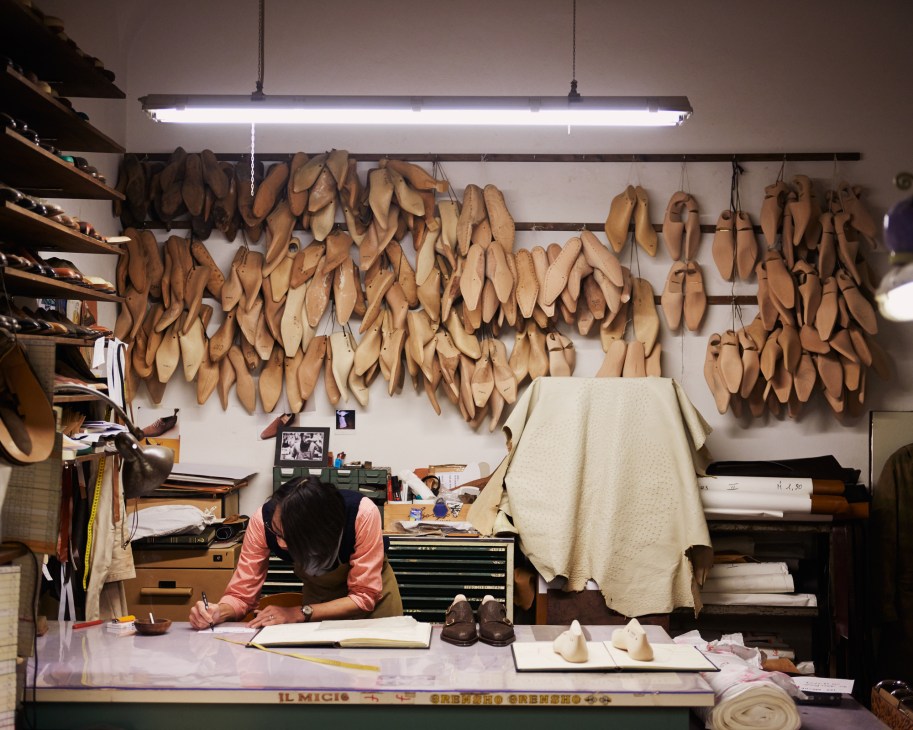 Shoemaker Hidetaka Fukaya sketching at his workbench beneath shoe lasts hung on the wall.