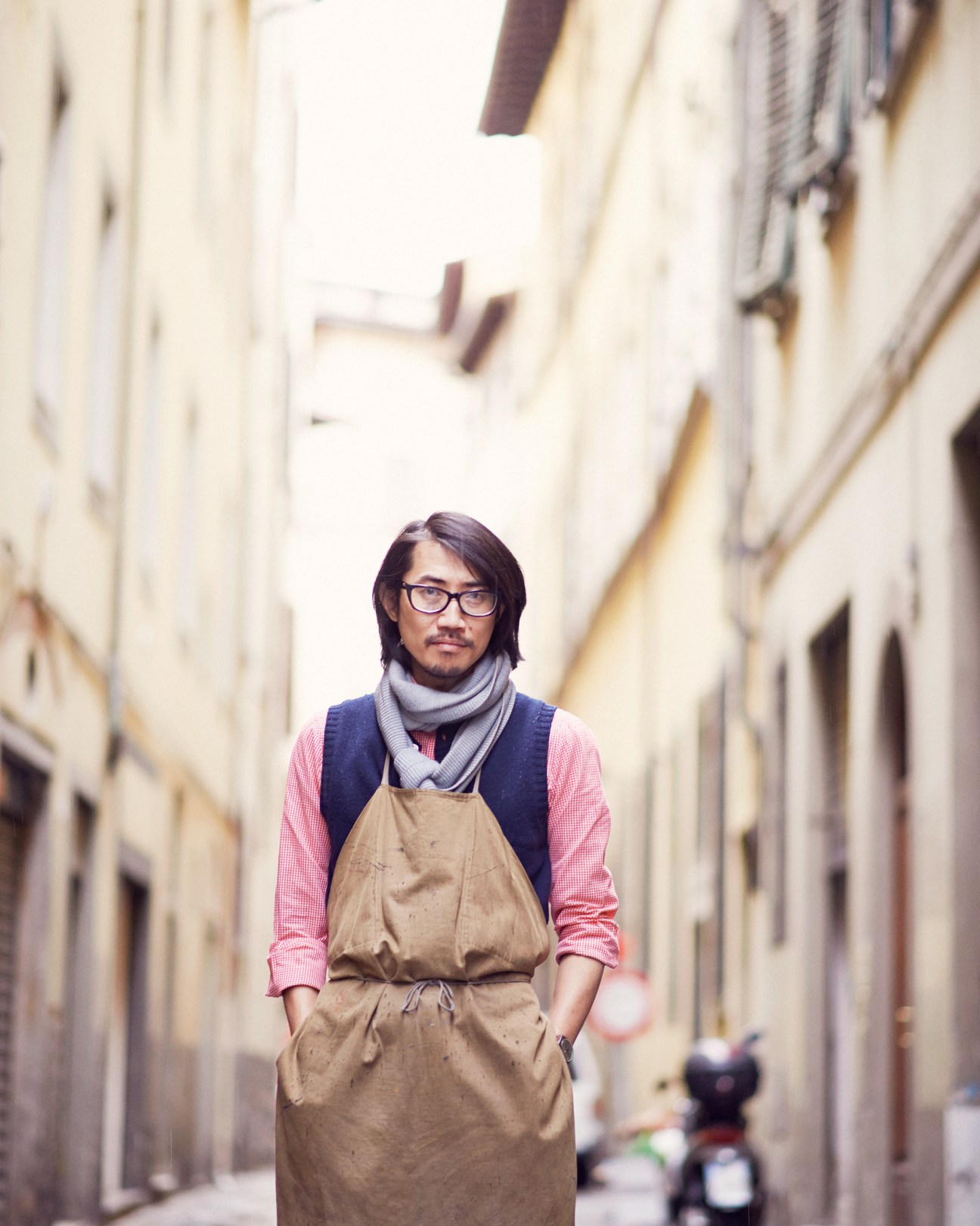 Hidetaka Fukaya standing in a narrow Florence street wearing glasses, a pink shirt, navy vest and tan apron.