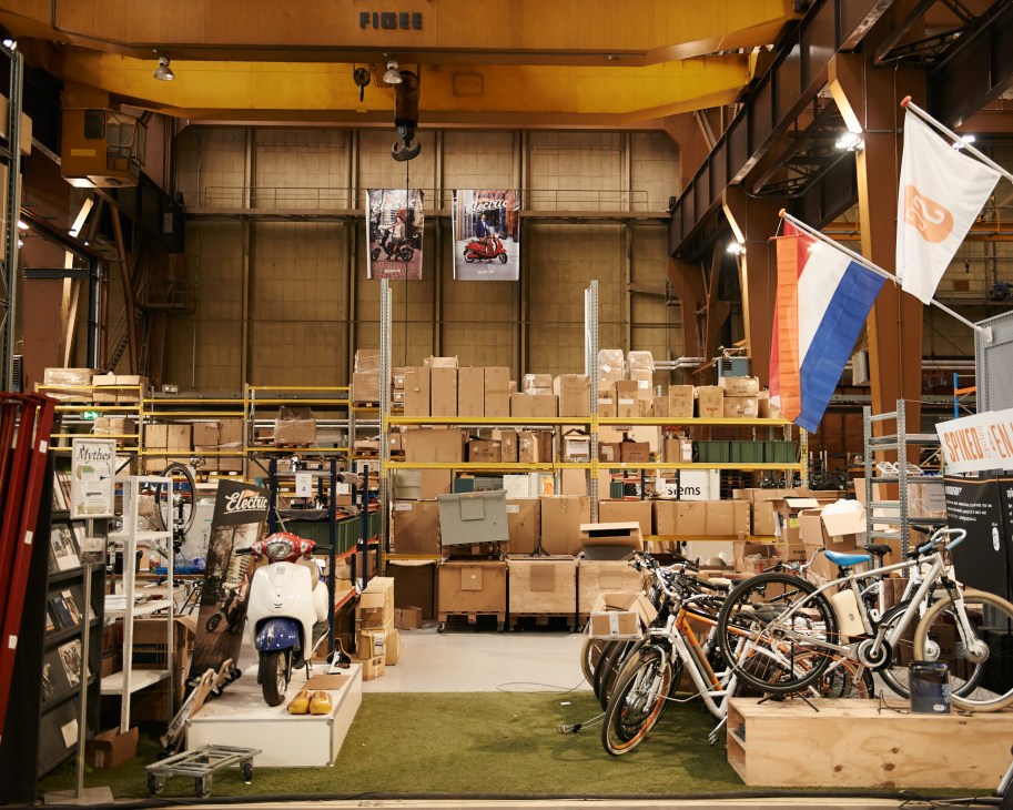 ESFA production studio in a former shipbuilding facility with yellow overhead cranes, cardboard boxes, assembled e-bikes and scooters displayed on the floor.