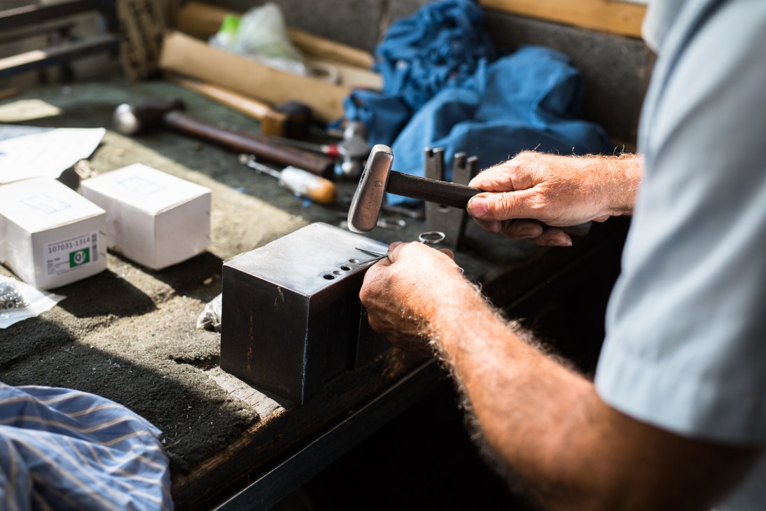 A craftsman using a steel hammer to make final adjustments to scissors on a metal block at a workbench.