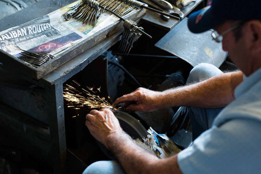 Craftsman Eric Stones polishes scissors on a grindstone, creating sparks, with finished scissors visible on the workbench behind him.