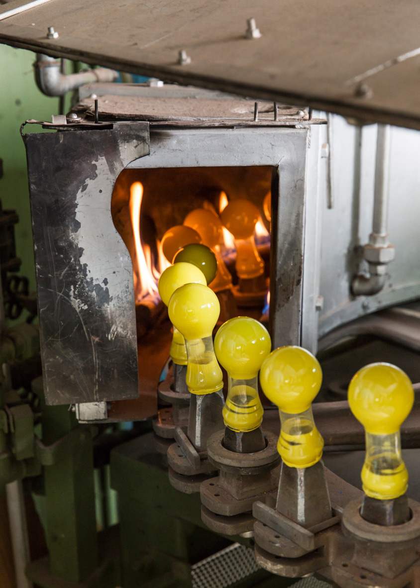 Yellow incandescent bulbs being heated on a carousel inside a metal chamber with open flame visible.