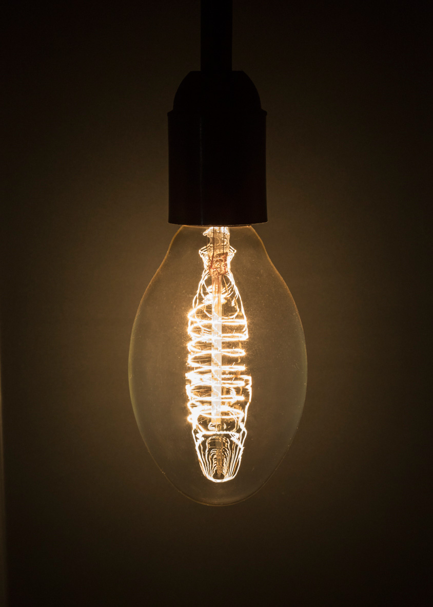 Illuminated incandescent bulb with intricate tungsten filament glowing warm amber against dark background.